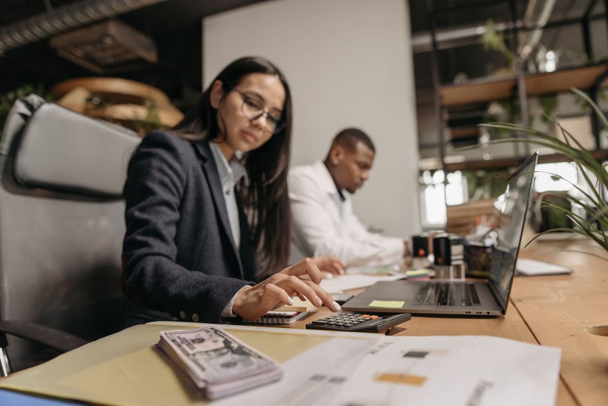 Office worker using a calculator at a desk covered in paperwork while a colleague works on a laptop