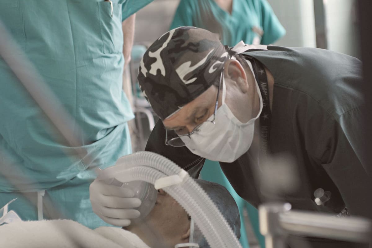 Clinician in a surgical cap and mask leaning over a patient while holding the breathing circuit in the operating room