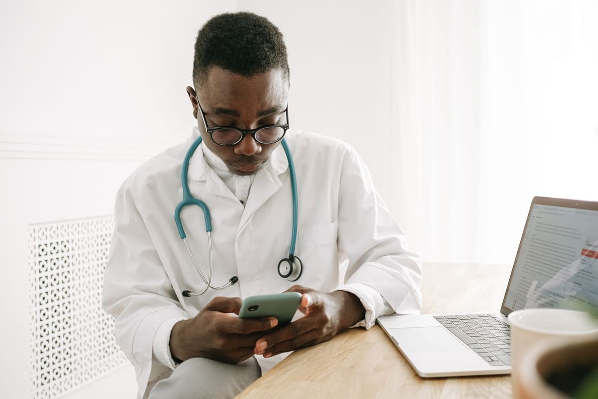 Doctor in a white coat using a smartphone at a desk beside an open laptop