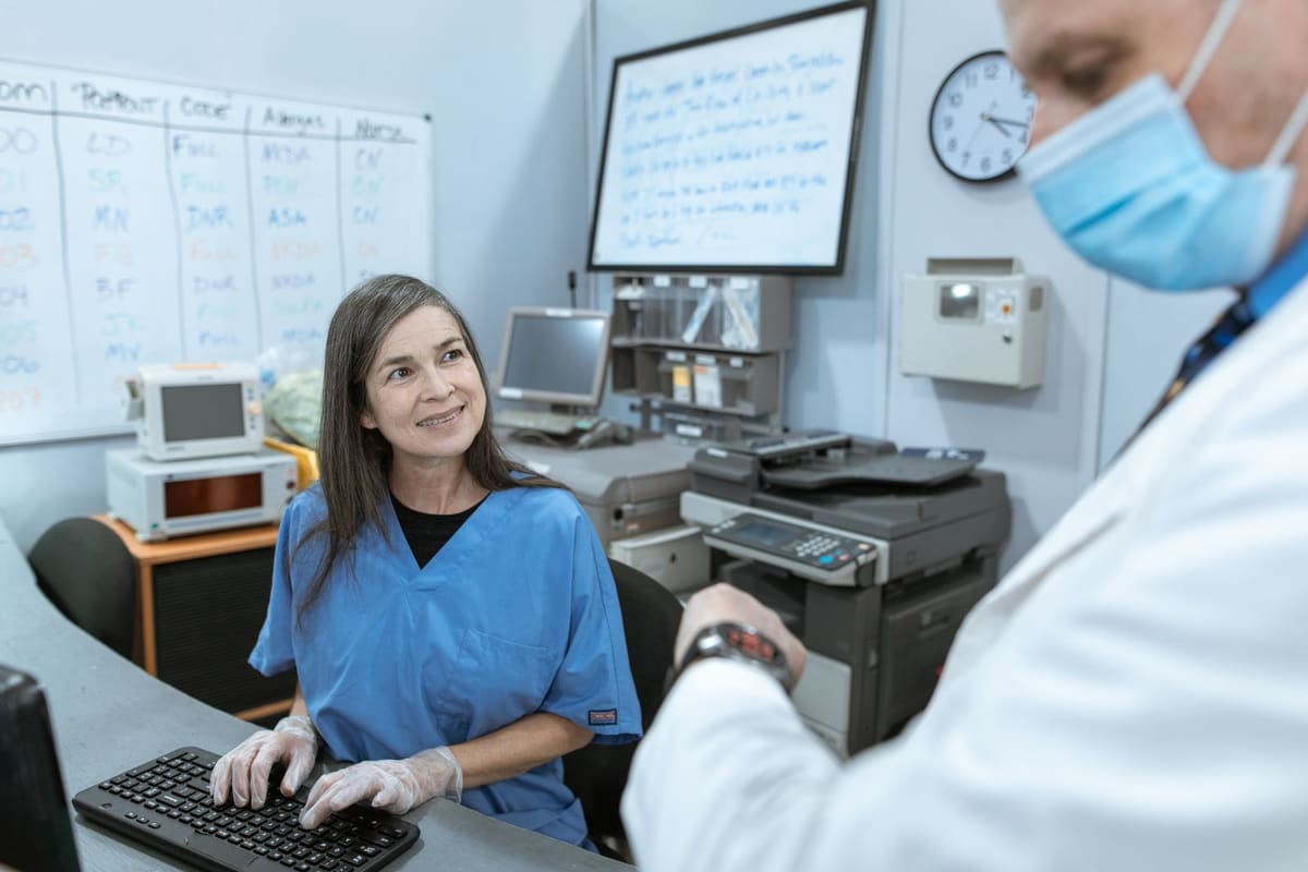 Medical office staff member in scrubs working at a computer while talking with a doctor