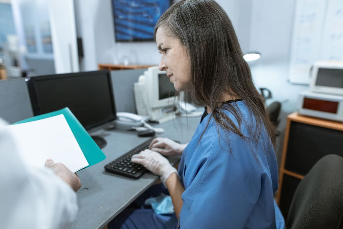 Clinician in blue scrubs and gloves typing at a computer workstation in a medical office