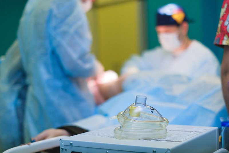 Anesthesia mask and equipment in the foreground while the surgical team works on a patient in the operating room
