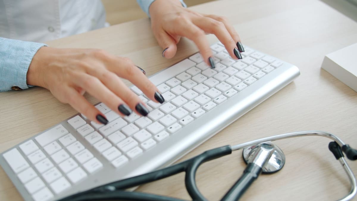 Hands typing on a computer keyboard beside a stethoscope